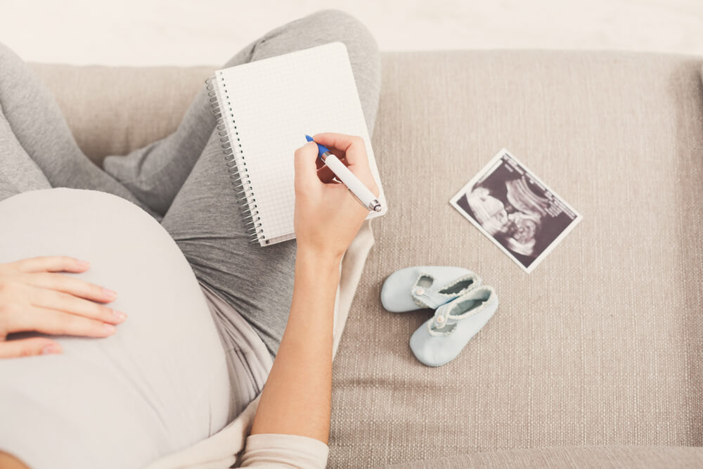 pregnant woman sitting on sofa with blank notebook writing names for her baby boy