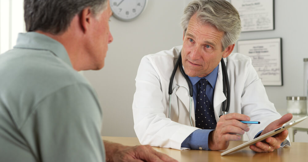 Mature Doctor Talking With Patient and Tablet in Office