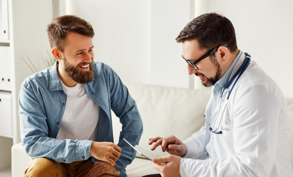 Young Bearded Guy Gesticulating and Talking With Friendly Male Doctor While Sitting on Sofa in Office of Modern Hospital