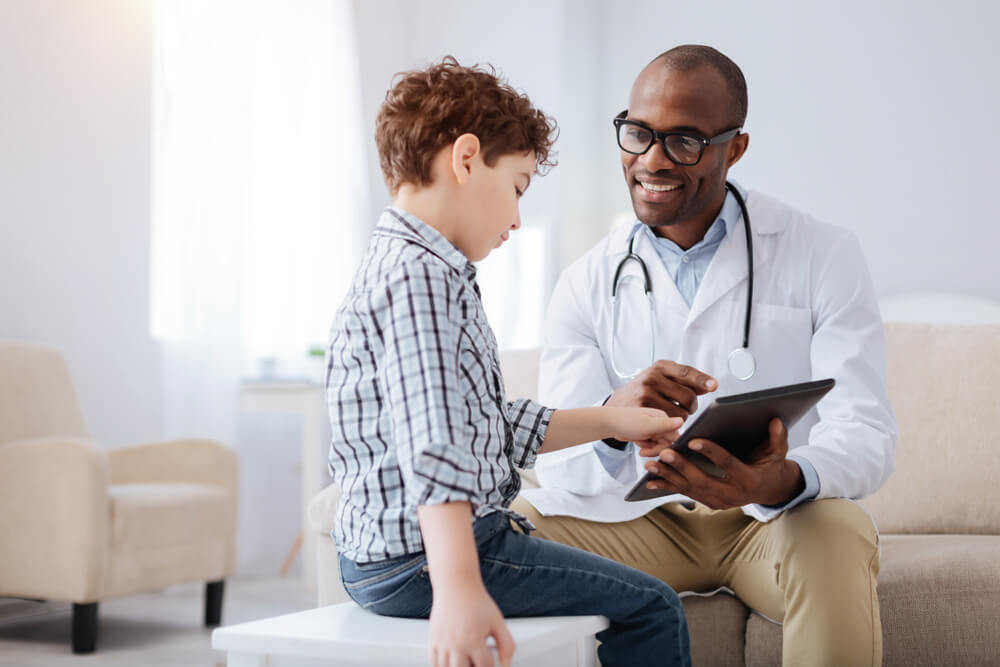 Positive Pleasant Male Doctor Smiling While Showing Tablet to Boy and Sitting