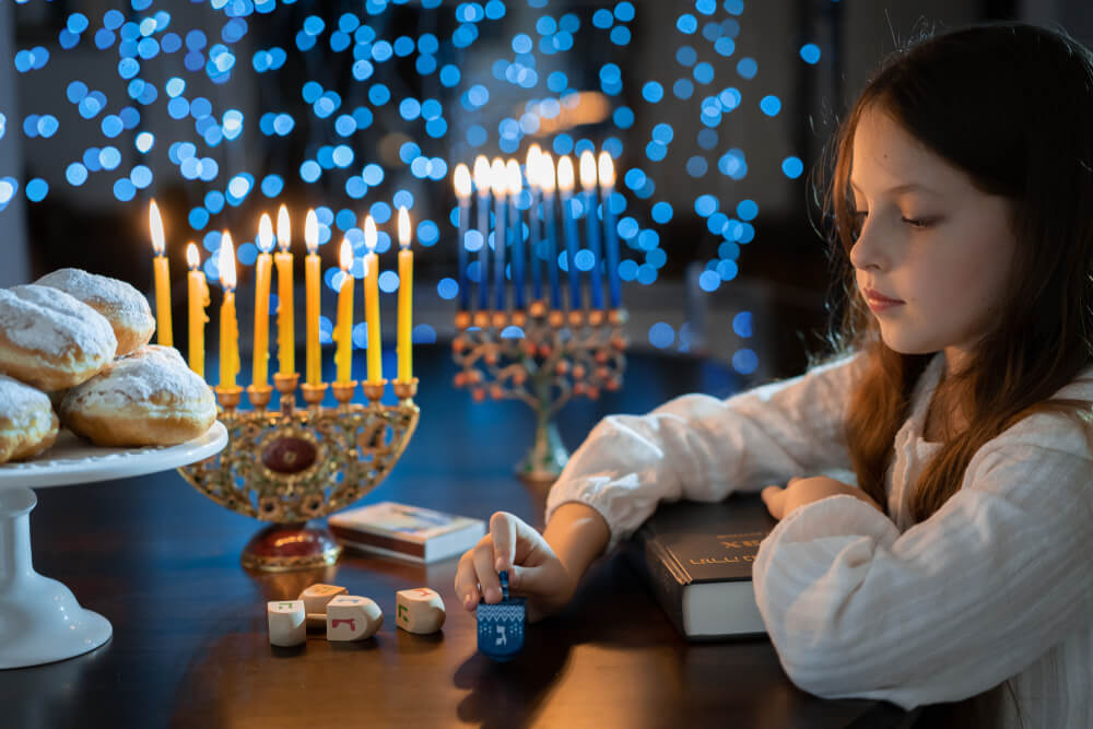 Child Girl Looking at Menorah Candles on Wooden Table and Sufganiyot on Background Light Glitter Bokeh Overlay.